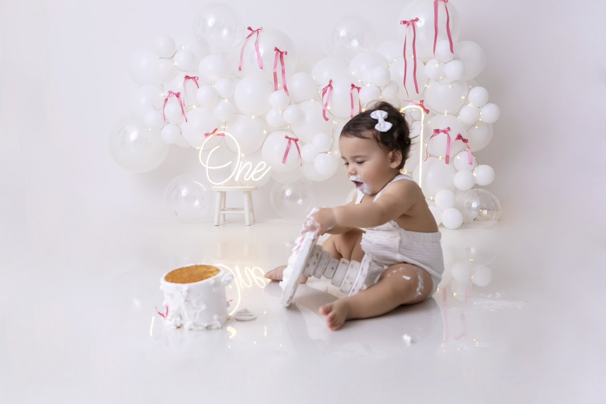 Bebê em ensaio Smash the Cake clean em estúdio branco com laços rosa, cenário minimalista e elegante, criança comendo e se lambuzando com bolo de aniversário em Caraguatatuba