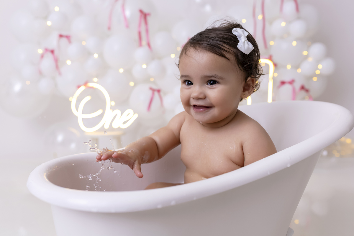Bebê em ensaio Smash the Cake clean em estúdio branco com laços rosa, cenário minimalista e elegante, criança comendo e se lambuzando com bolo de aniversário em Caraguatatuba