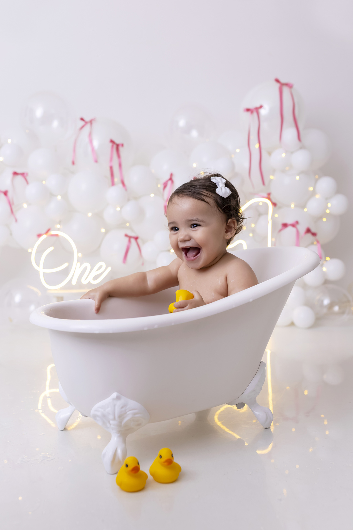 Bebê em ensaio Smash the Cake clean em estúdio branco com laços rosa, cenário minimalista e elegante, criança comendo e se lambuzando com bolo de aniversário em Caraguatatuba