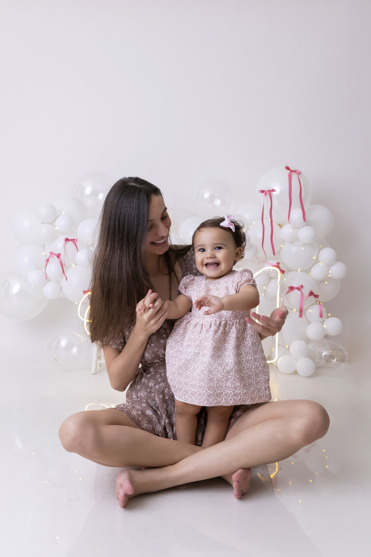 Bebê em ensaio Smash the Cake clean em estúdio branco com laços rosa, cenário minimalista e elegante, criança comendo e se lambuzando com bolo de aniversário em Caraguatatuba