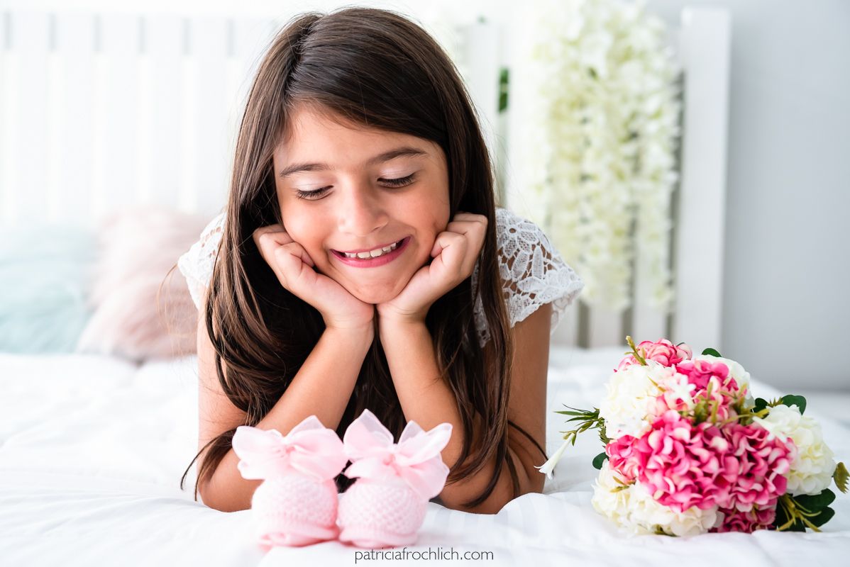Menina criança deitada sorrindo vestindo branco e olhando sapatinhos rosa de bebe. Ensaio fotografico com luz natural em estudio na Gloria Zona Sul do Rio de Janeiro 