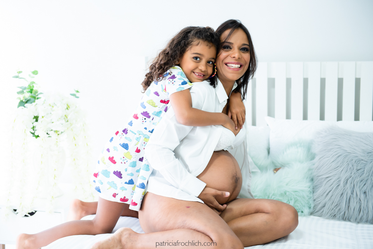 Ensaio fotografico familia gestante. Mãe e filha abraçadas na cama, gestante com a mão na barriga de blusa branca. Ensaio em estudio na Gloria Zona Sul do Rio de Janeiro com luz natural 