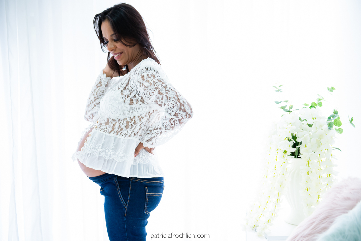 Ensaio gestante com blusa branca e calça jeans fazendo pose de lado. Ensaio em estudio na Gloria Zona Sul do Rio de Janeiro com luz natural 