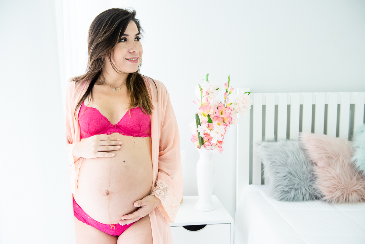 Gestante com lingerie rosa de pé ao lado de janela e proxima da cama e do criado mudo em ensaio gestante gravida em estúdio com luz natural