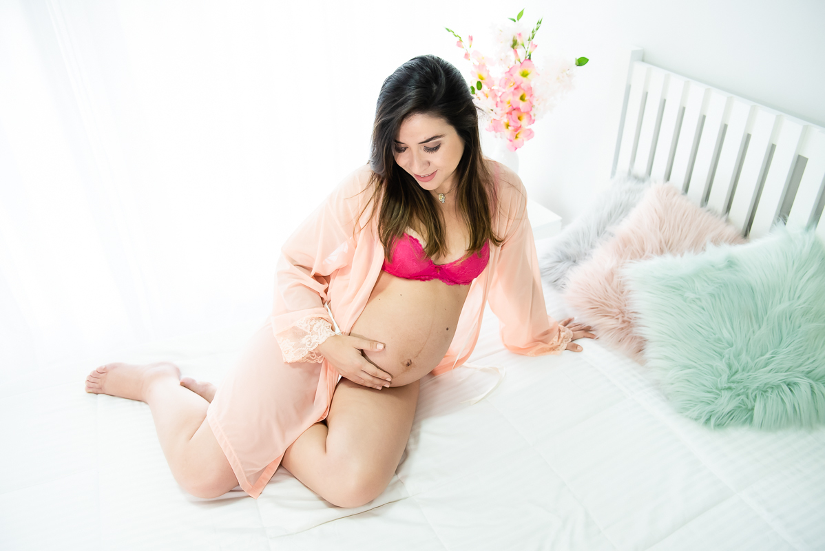 gestante fazendo pose com a mão na barriga sobre a cama branca em ensaio gestante gravida em estúdio com luz natural