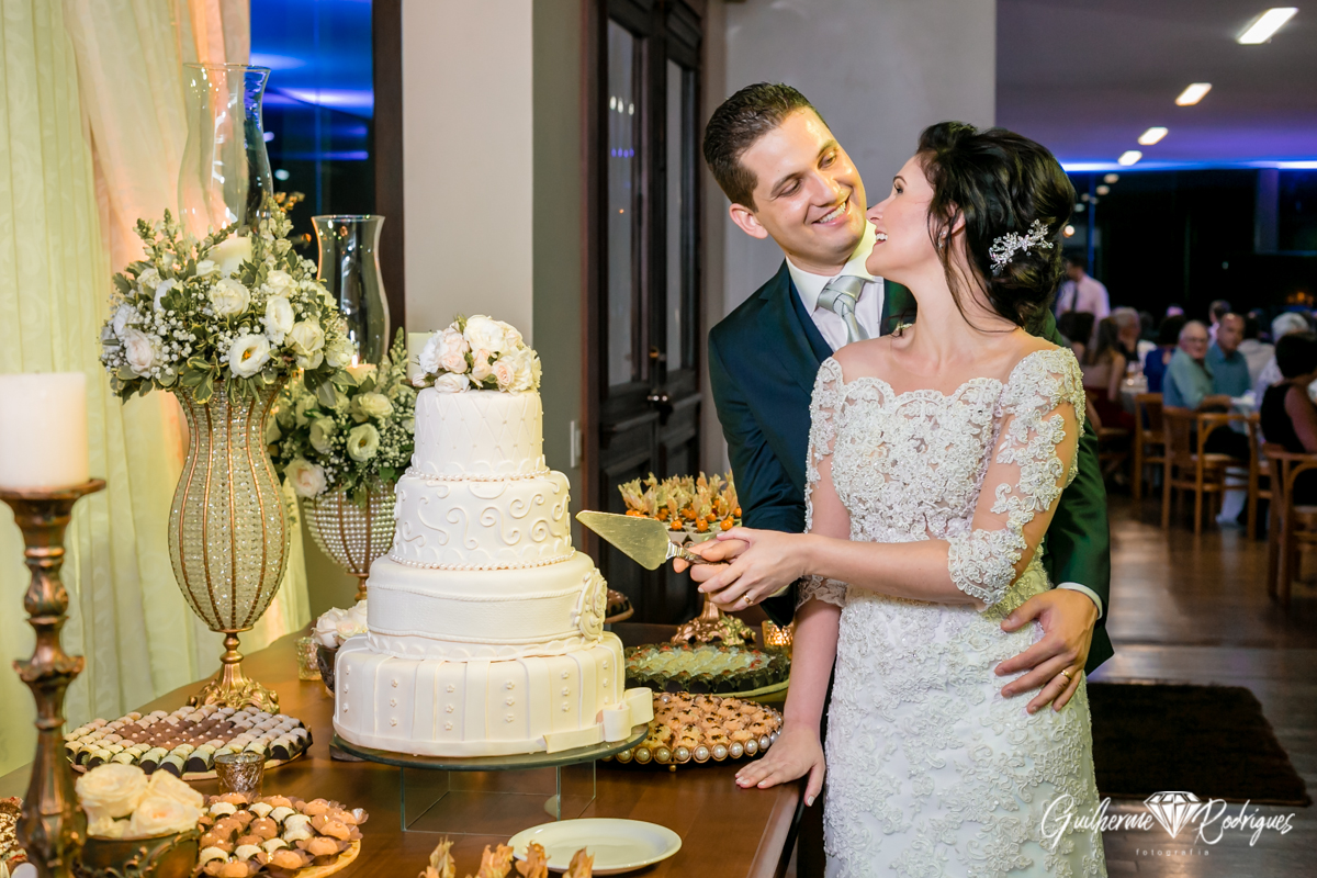 Corte do bolo de casamento, noivo abraçado com a noiva para fazer o corte do bolo. Registro fotográfico Guilherme Rodrigues.