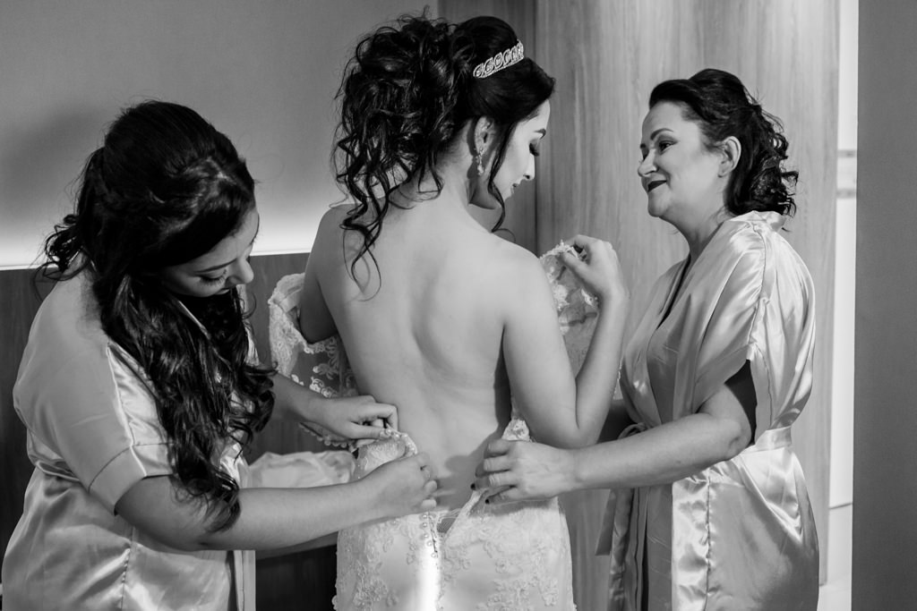 A noiva Rochele recebendo a ajuda da sua mãe e sua irmã vestindo o vestido