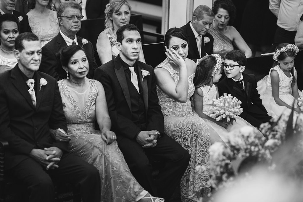 os pais da noiva com olhar firme para a benção do padre e os irmãos da noiva emocionados e a troca de olhares do pagem com as daminhas.