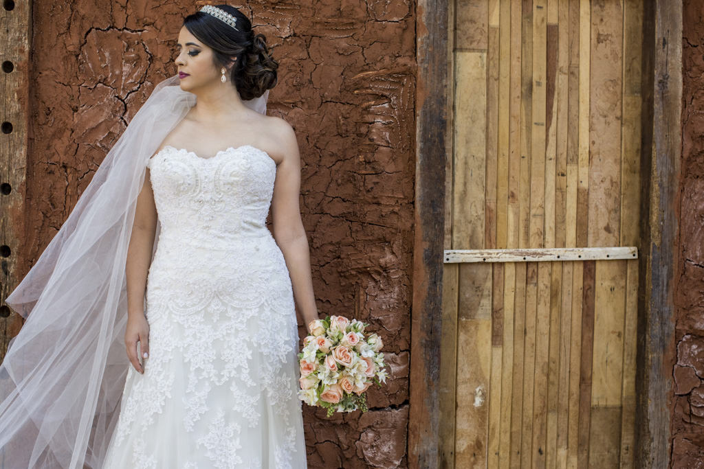 Toda a elegância de thammy com seu lindo vestido de noiva segurando o buquê ao fundo um parede de pau a pique