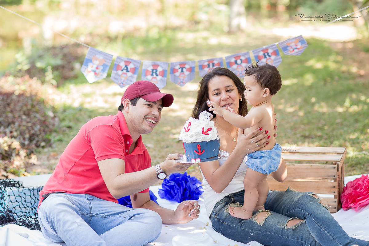 O papai levantando o bolo e colocando perto do Arthur porque ele esta com um pouco de receio de se sujar com o bolo
