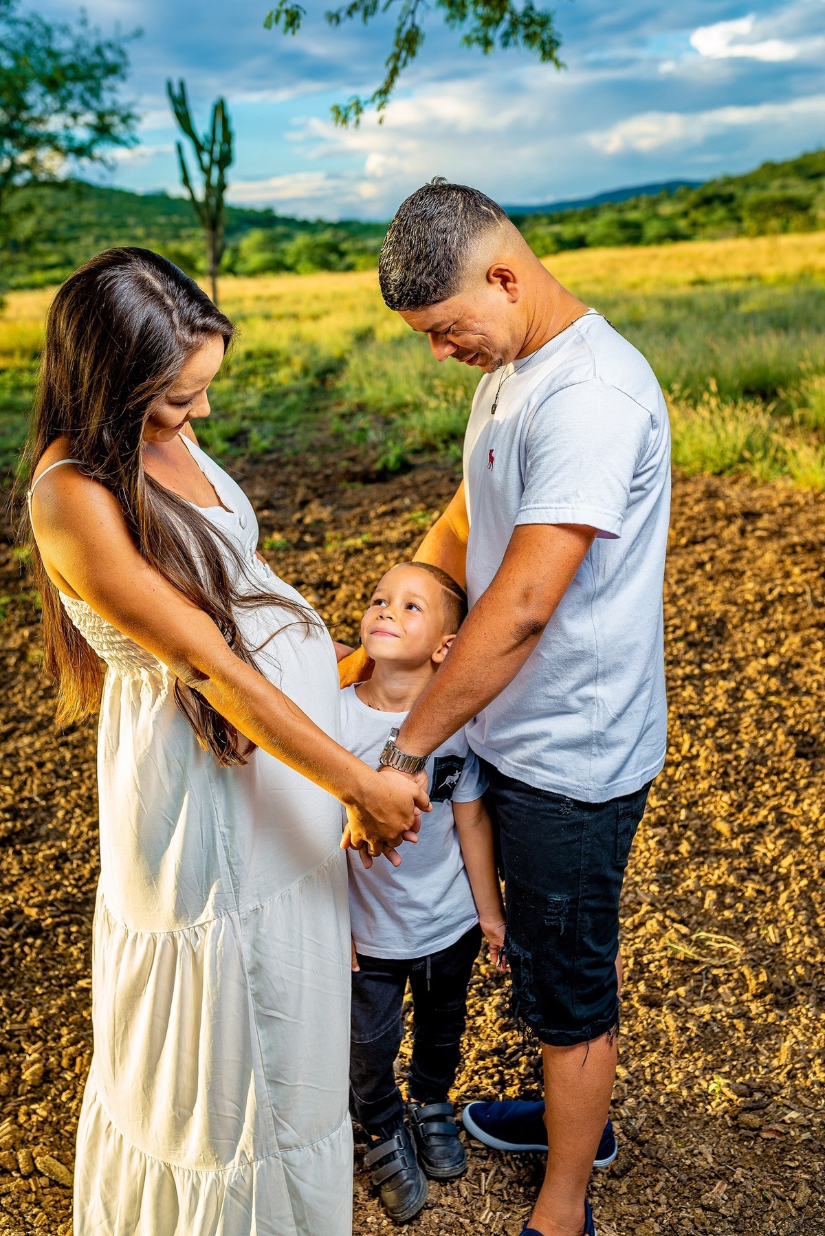 mãe gestante e e pai de mãos dadas com o filho mais velho no meio dos dois olhando para cima e sorrindo