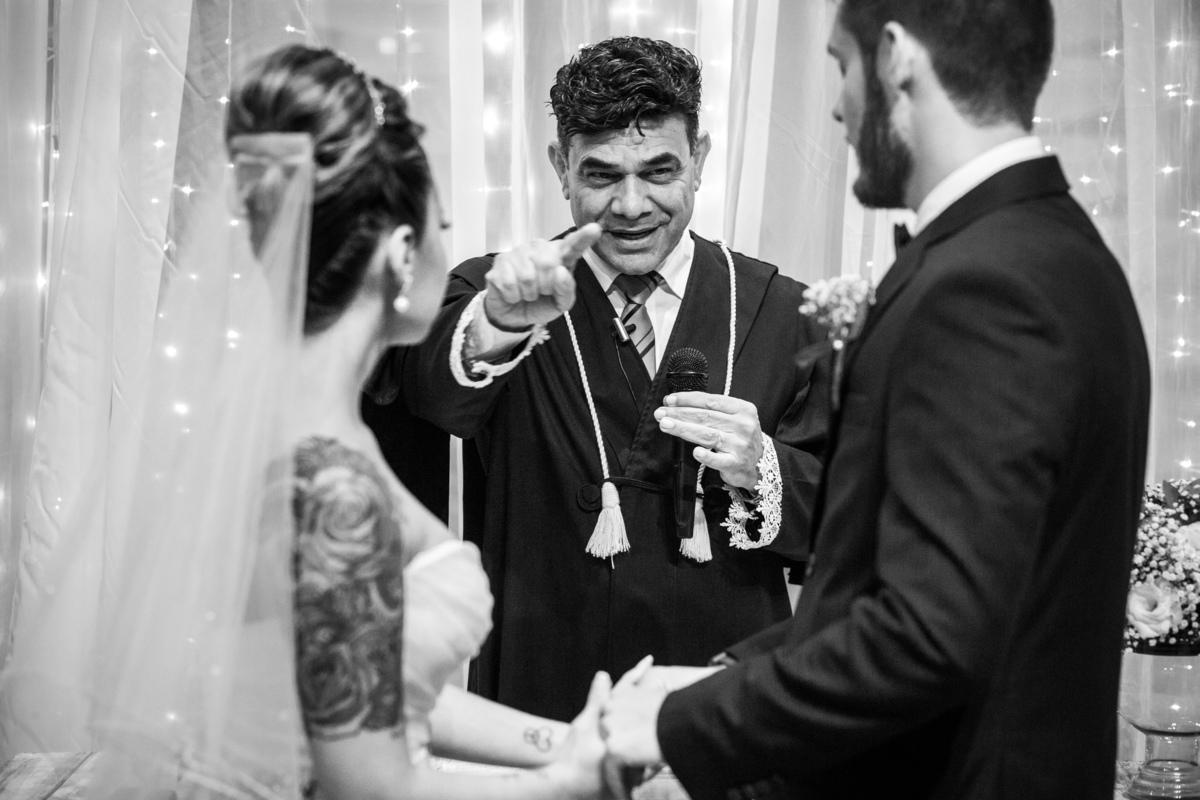 padre sorri apontando noivos no altar observam noiva tatuada preto e branco
