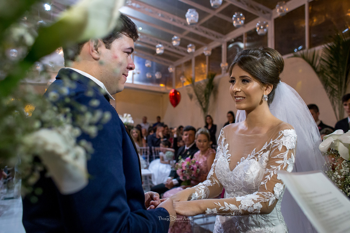 casamento cerimônia noivos ouvem o pastor decoração flores copo de leite palmeiras luminárias no teto convidados ao fundo