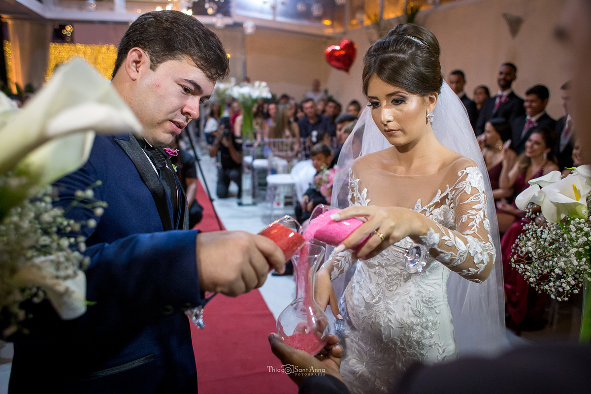 noivos despejam líquidos no mesmo recipiente decoração flores copo de leite convidados ao fundo tapete vermelho  