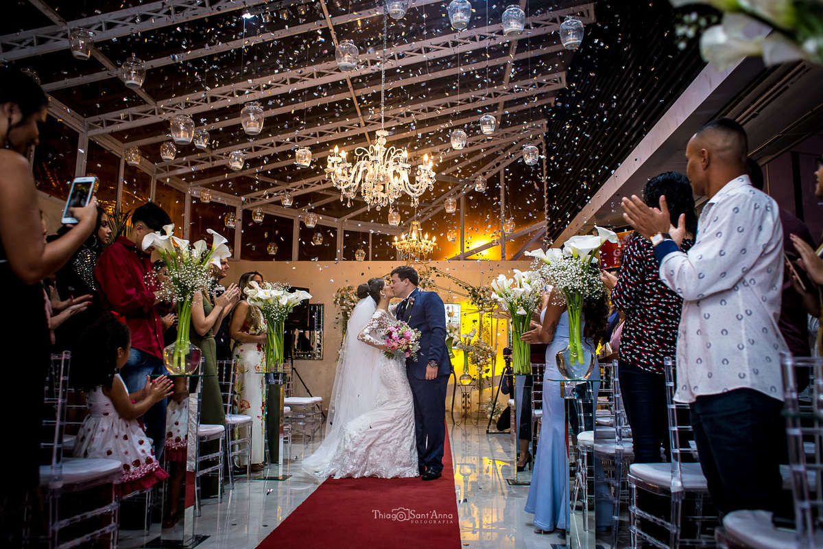 convidados aplaudem beijo dos noivos tapete vermelho decoração luminárias no teto flores copo de leite 