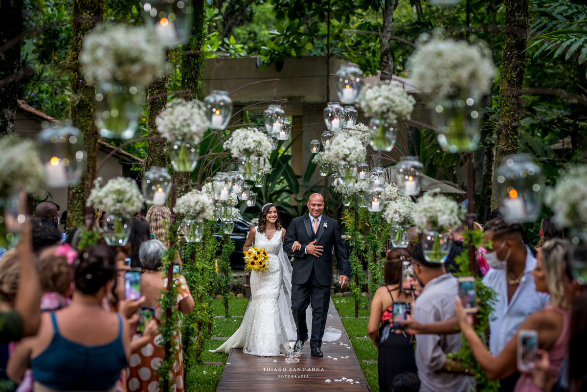 entrada da noiva com seu pai convidados observam de pé ambiente externo árvores gramado decoração jarros flores brancas