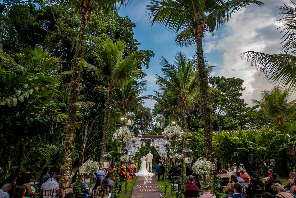 noivos no altar convidados observam ambiente externo decoração arranjos flores brancas vegetação palmeiras