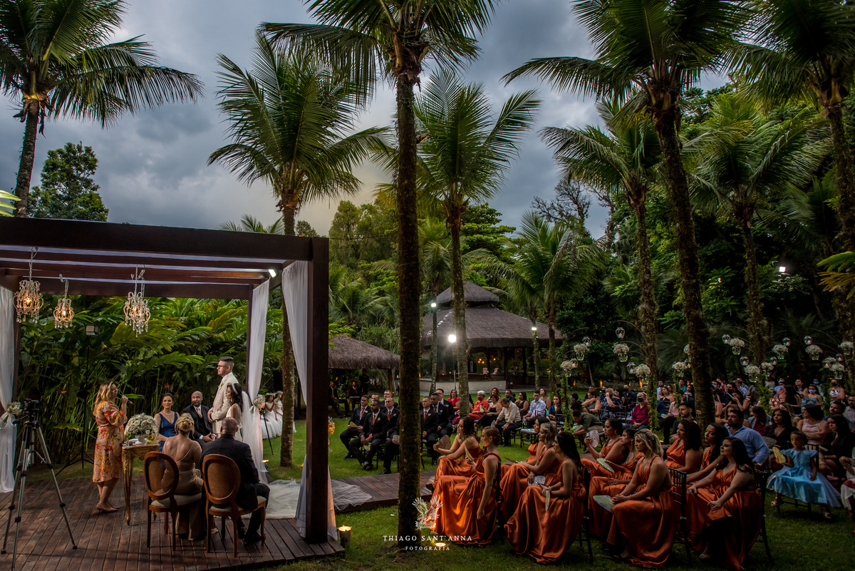 noivos altar estrutura de madeira tecidos brancos padrinhos e convidados observam ambiente externo vegetação gramado palmeiras flores brancas