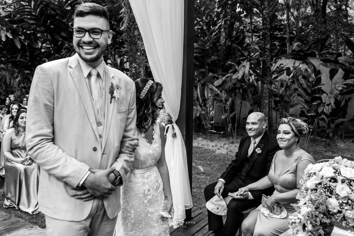 noivos sorrindo altar junto a familiares ambiente externo vegetação ao fundo decoração flores preto e branco