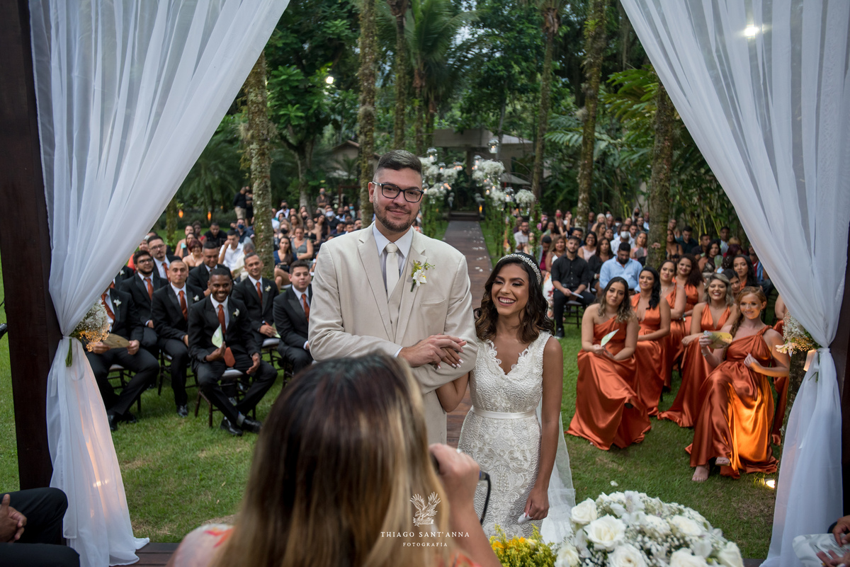 noivos no altar sorrindo atentos a pastora cortinas tecido branco padrinhos convidados ao fundo vegetação gramado palmeiras 