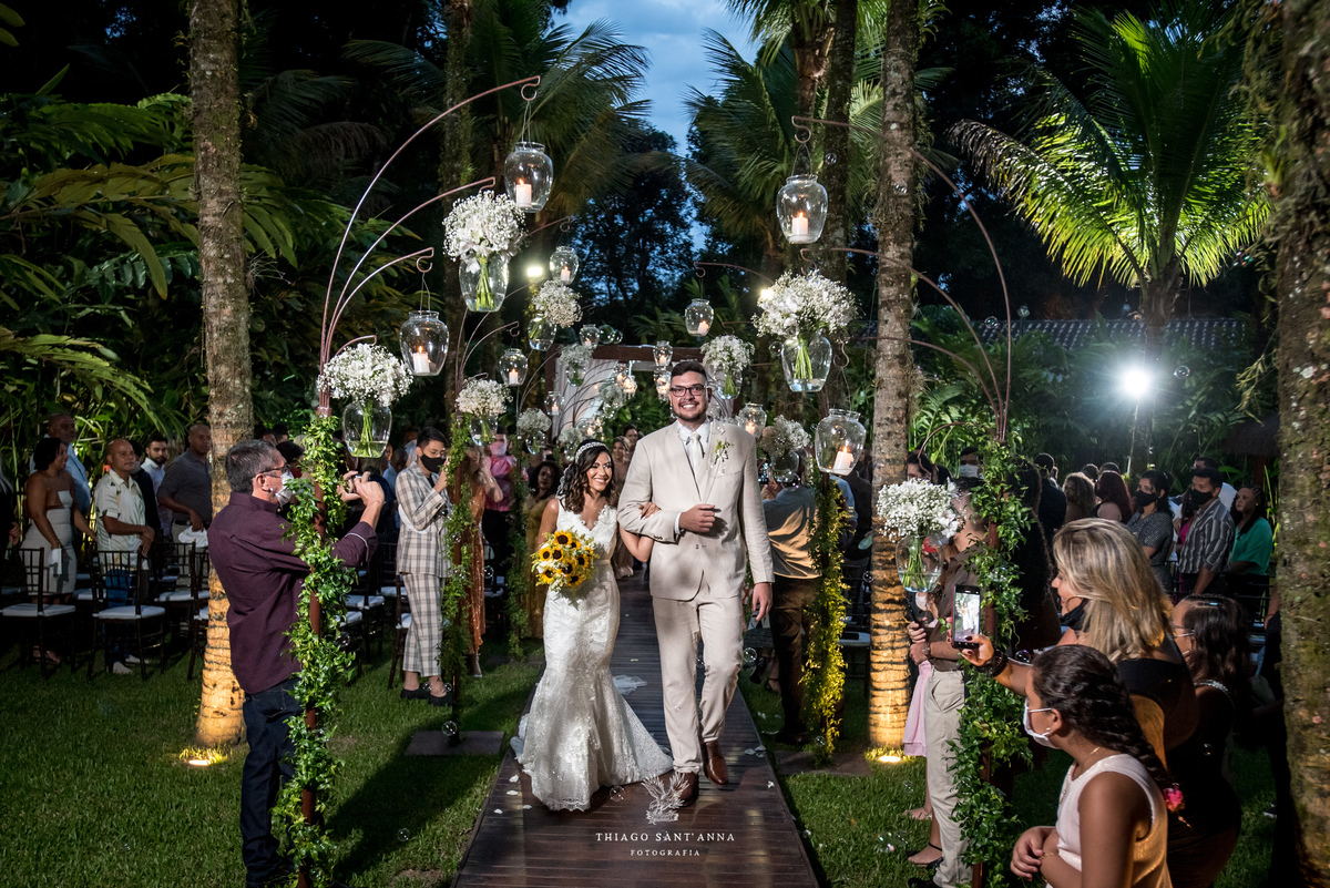 saída do casal sorriso noiva vestido buque girassol noivo terno bege ambiente externo vegetação arranjos flores brancas