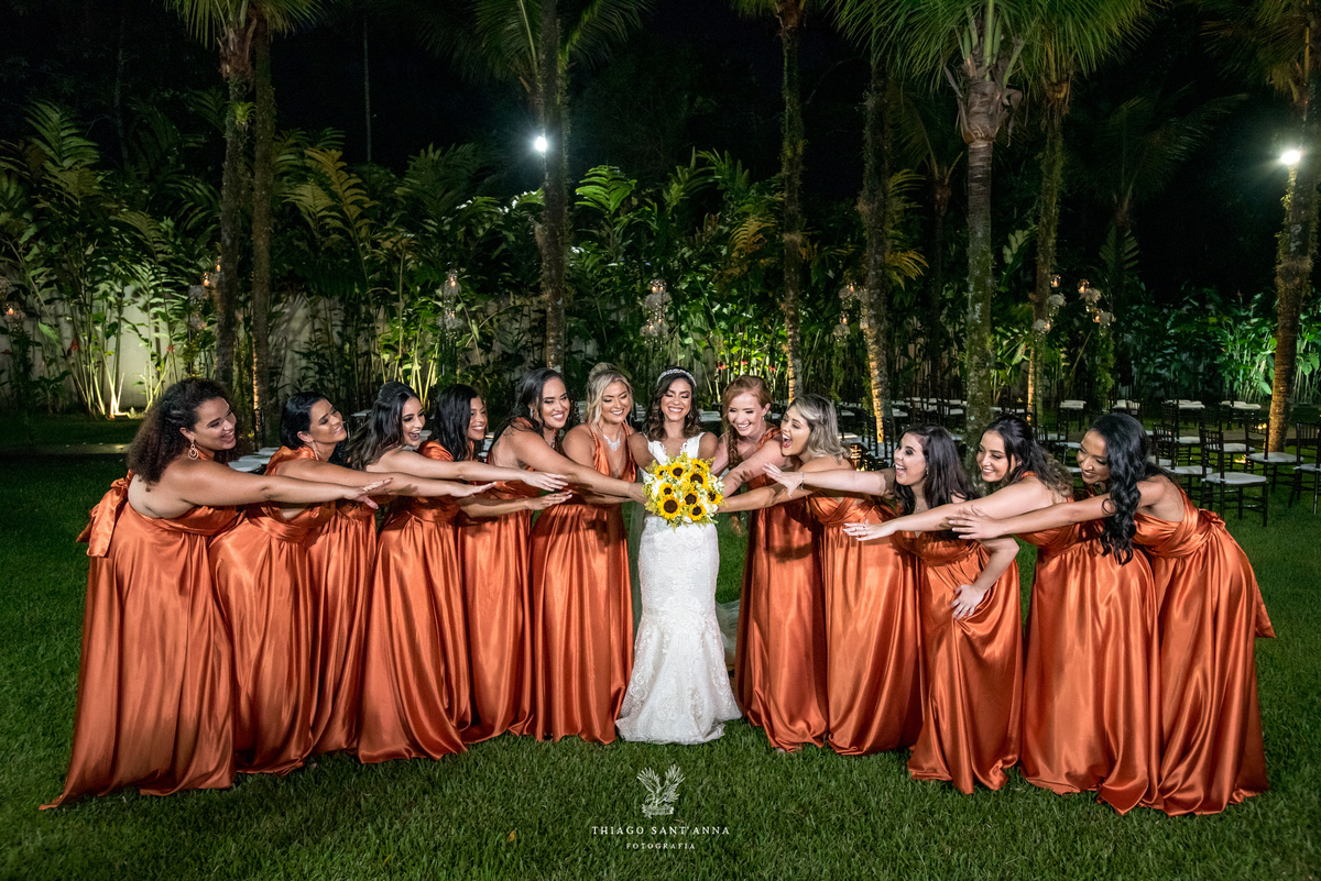 noiva posa com madrinhas vestido laranja cobre estendem as mãos em direção ao buque ambiente externo vegetação palmeiras gramado