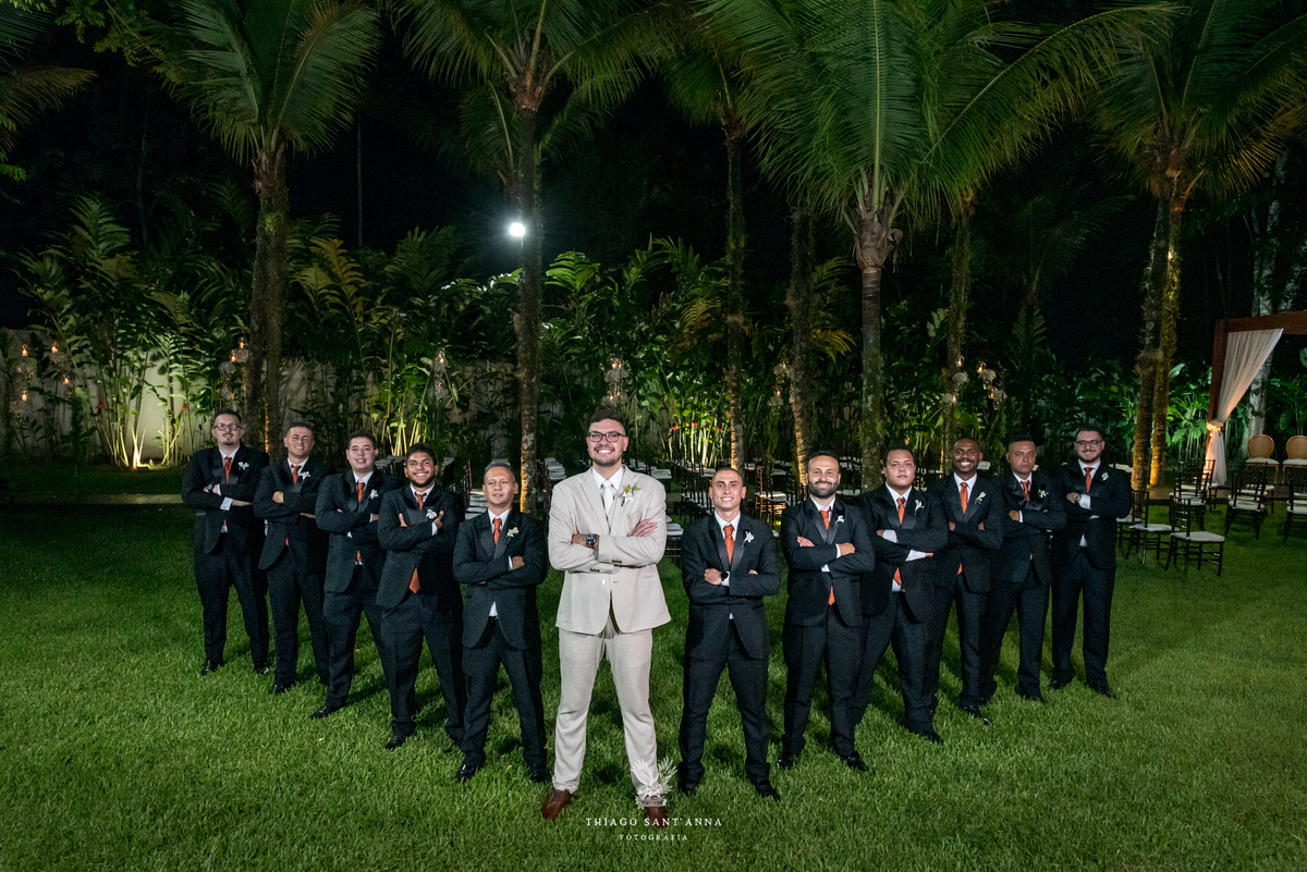 noivo terno bege posa com padrinhos de braços cruzados terno preto ambiente externo vegetação palmeiras gramado