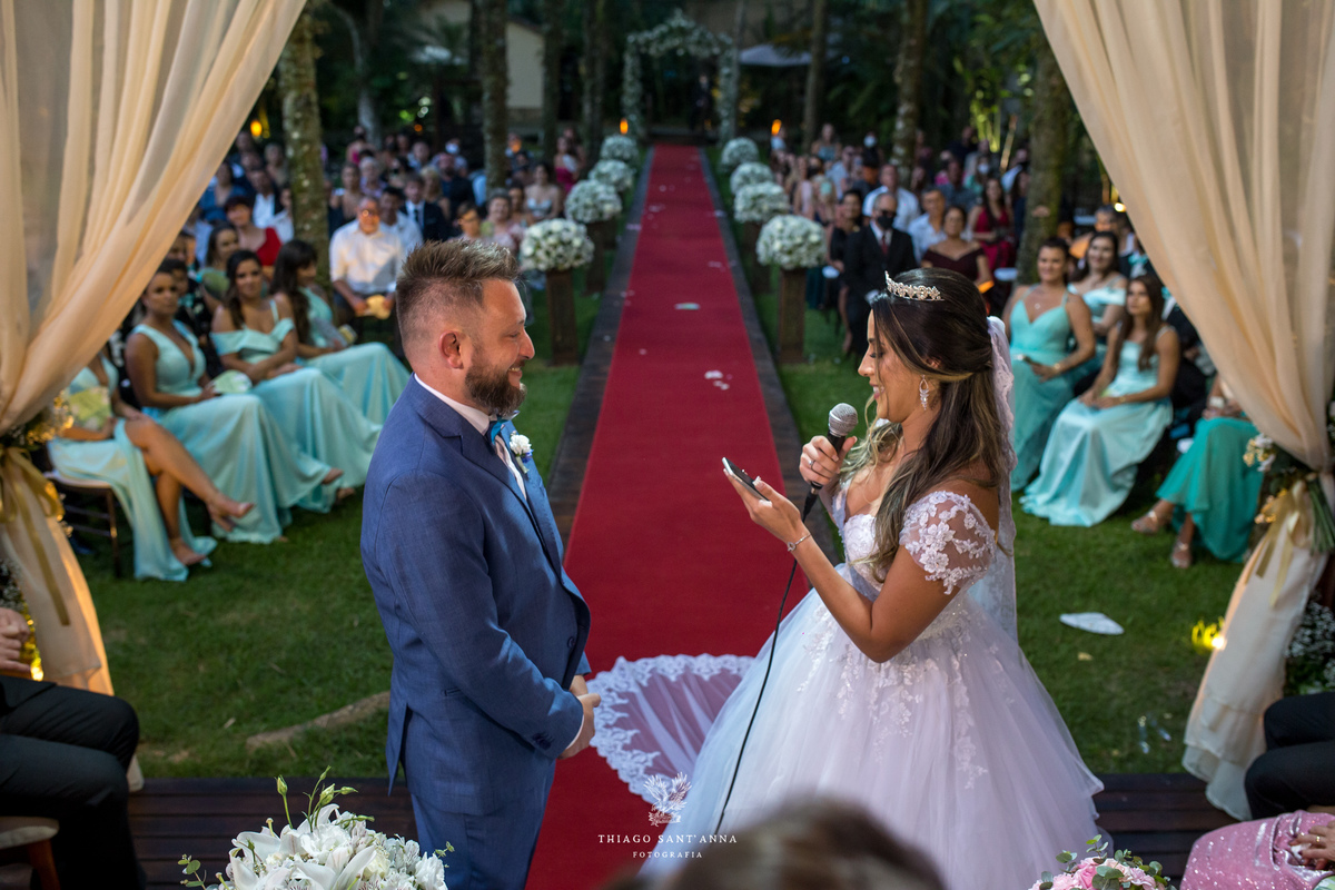 noivos no altar noiva lê seus votos madrinhas e convidados ao fundo tapete vermelho decoração flores brancas
