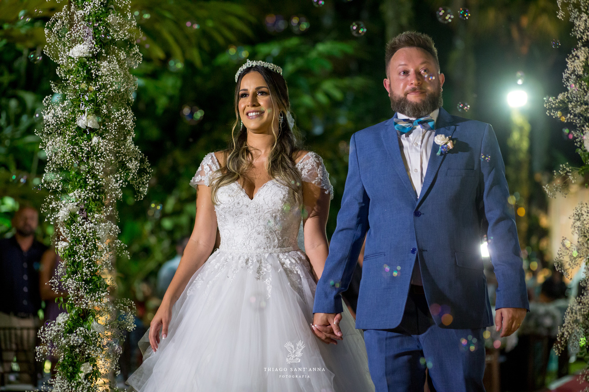 noivos de mãos dadas bolas de sabão ambiente externo vegetação flores brancas noivo terno azul noiva vestido renda 