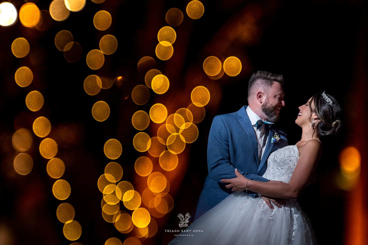 noivos sorrindo se observam  luzes laranja ao fundo