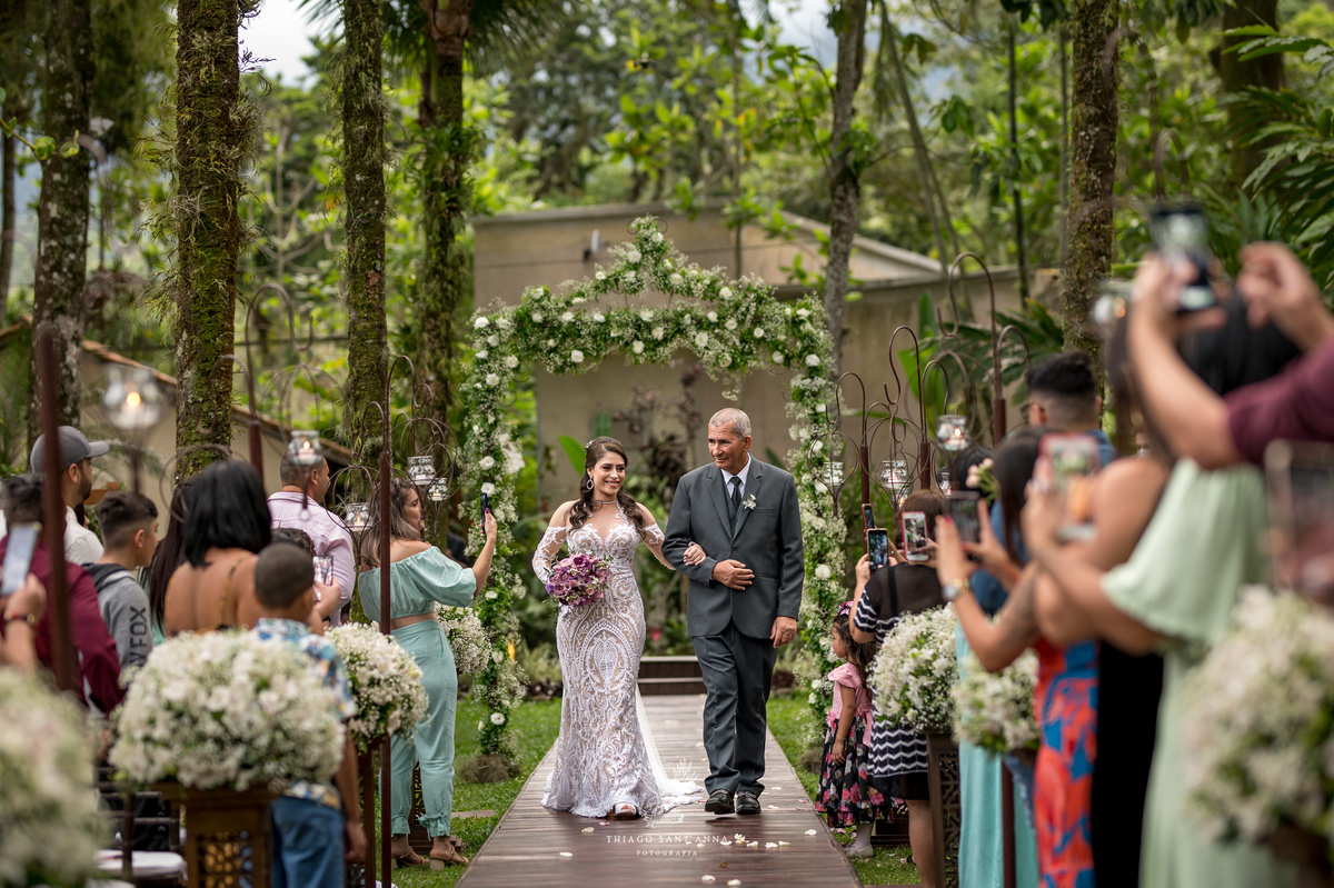 entrada noiva e seu pai convidados observam de pé decoração flores brancas ambiente externo vegetação gramado coqueiros