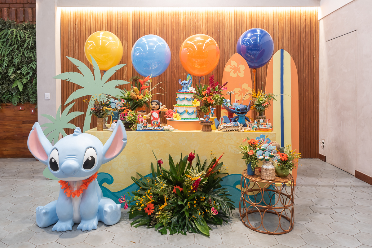 Decoração de festa infantil tema Lilo e Stitch com mesa principal colorida, balões, prancha de surf, bolo decorado e personagem Stitch em destaque, em buffet infantil.
