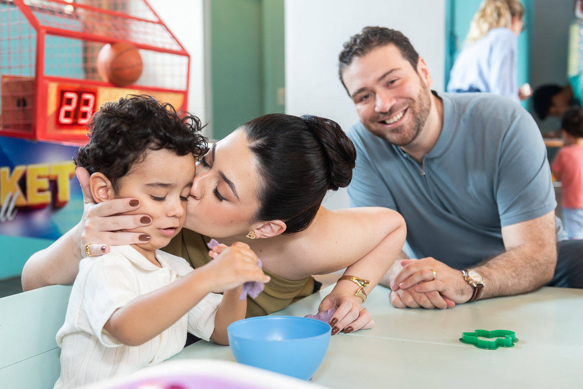 Mãe beijando filho durante festa infantil em buffet, com pai sorrindo ao fundo, momento de carinho e família em aniversário infantil.