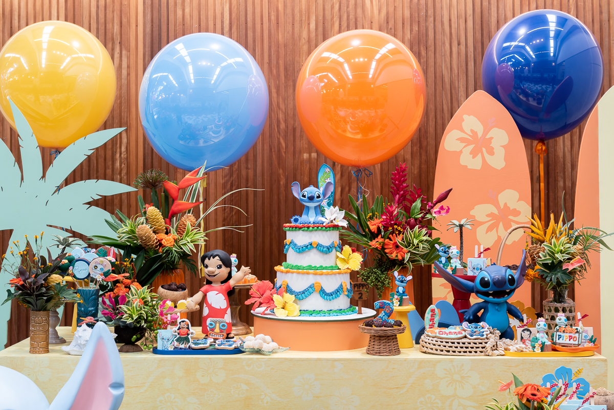 Decoração de festa infantil tema Lilo e Stitch com mesa principal colorida, balões, prancha de surf, bolo decorado e personagem Stitch em destaque, em buffet infantil.