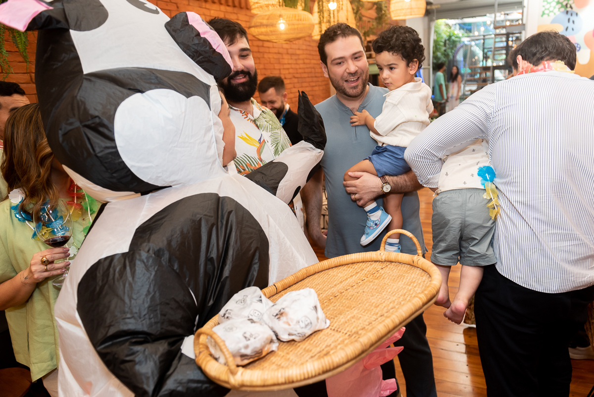 Personagem fantasiado de vaquinha servindo hambúrguer para convidados durante festa infantil em buffet, com família e crianças interagindo em ambiente animado de aniversário infantil na Casa Sapoti.