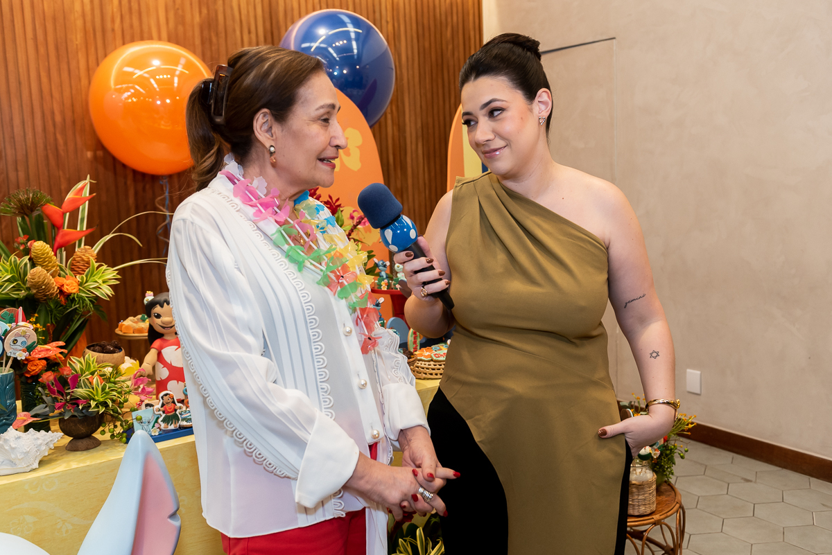Mulher entrevistando convidada com microfone durante festa infantil tema Lilo e Stitch, com decoração colorida, balões e mesa principal ao fundo em buffet infantil.

