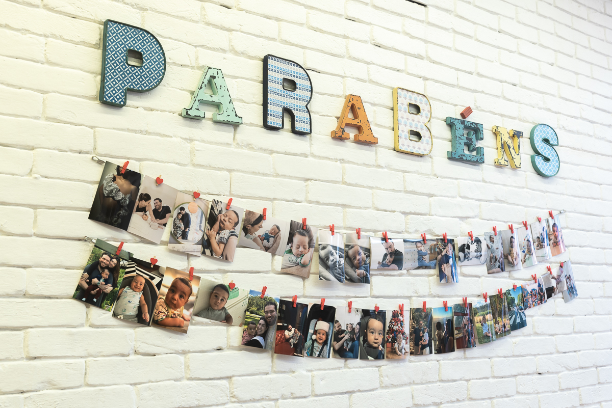 Decoração de festa infantil com painel de fotos do bebê em varal e letras coloridas “Parabéns” em parede branca, registro de aniversário infantil em buffet.
