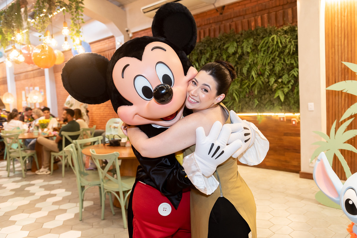 Mulher abraçando personagem Mickey Mouse durante festa infantil em buffet, momento espontâneo com decoração temática e convidados ao fundo.