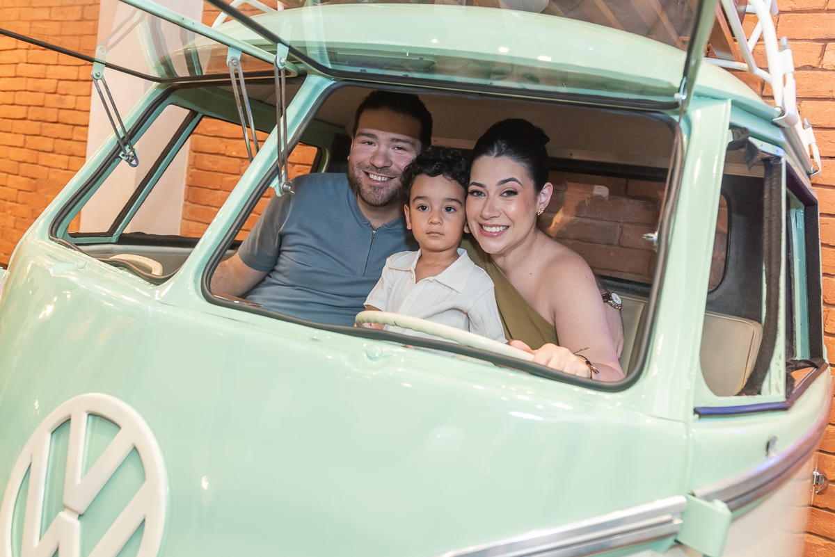 Família sorrindo dentro de kombi vintage decorativa durante festa infantil em buffet, pais e criança posando para foto em ambiente temático.