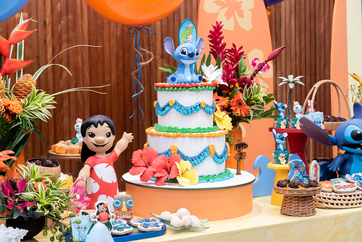 Decoração de festa infantil tema Lilo e Stitch com mesa principal colorida, balões, prancha de surf, bolo decorado e personagem Stitch em destaque, em buffet infantil.