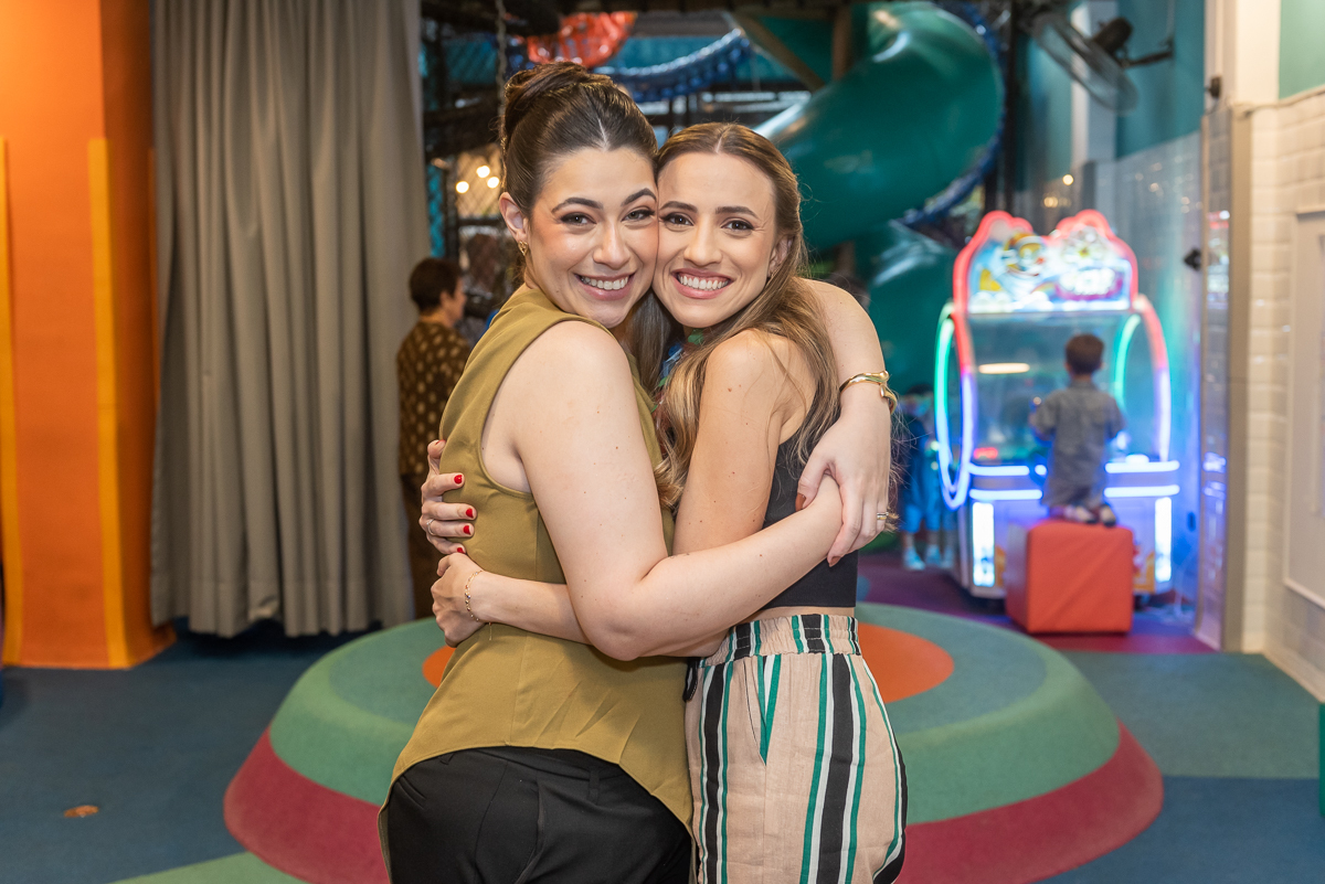 Duas mulheres sorrindo e se abraçando durante festa infantil em buffet, com área de brinquedos e fliperama ao fundo, momento de alegria entre convidados.