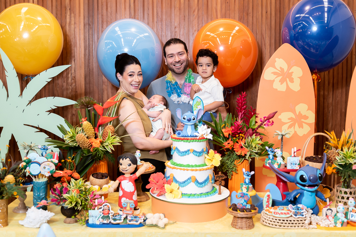 Família reunida na mesa do bolo em festa infantil tema Lilo e Stitch, com decoração tropical, balões coloridos e bolo personalizado em buffet infantil.