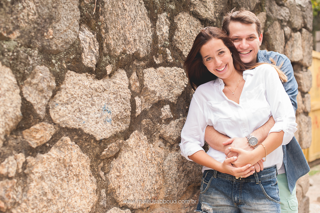 foto de casal. marido atrás da noiva e abraçando ela pela cintura. Ambos estão sorrindo e encostados em um muro de pedra.