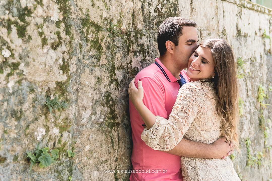 foto, ensaio de casal, um de frente para o outro com o noivo encostado no muro de pedras e dando um beijo no rosto da noiva.
