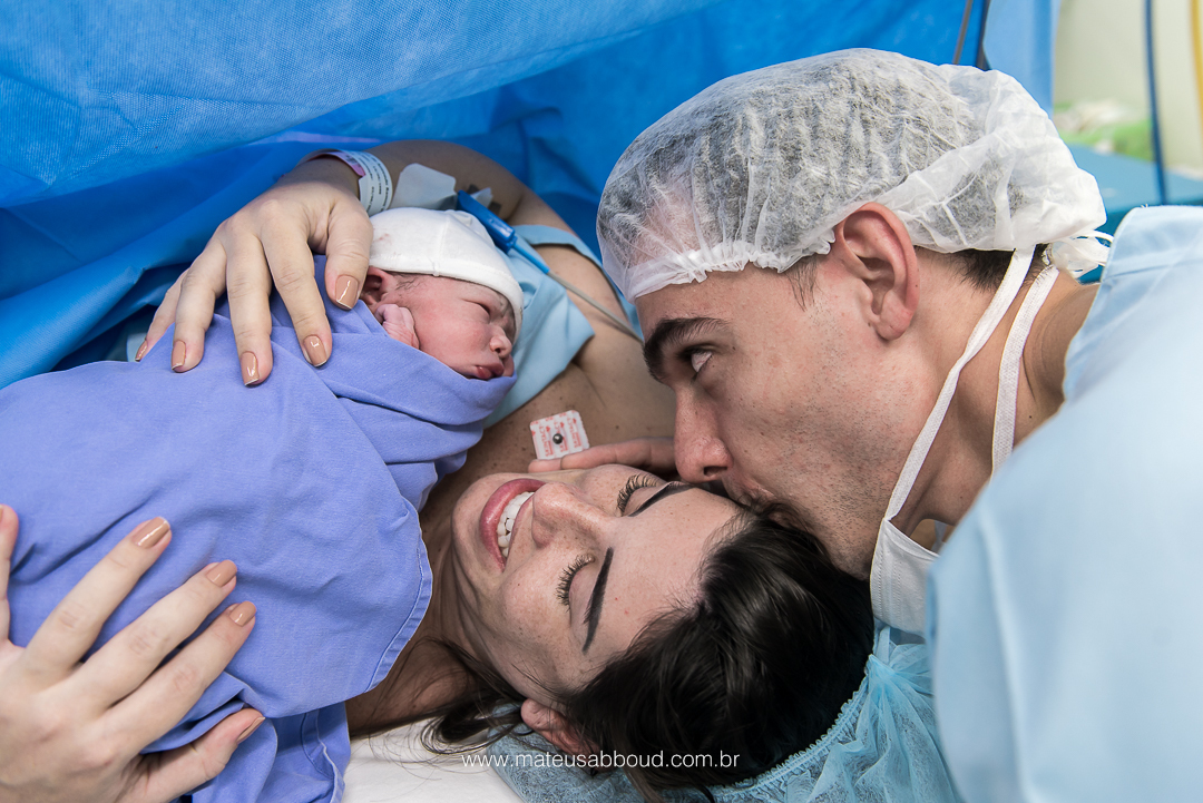 Pai beijando a cebeça da mamãe enquanto ela segura o bebê nos braços.