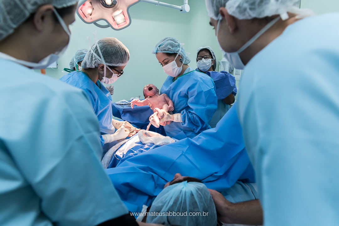 Bebê já fora da barriga nos braços da obstetra. Mãe e pai olham atentamente.