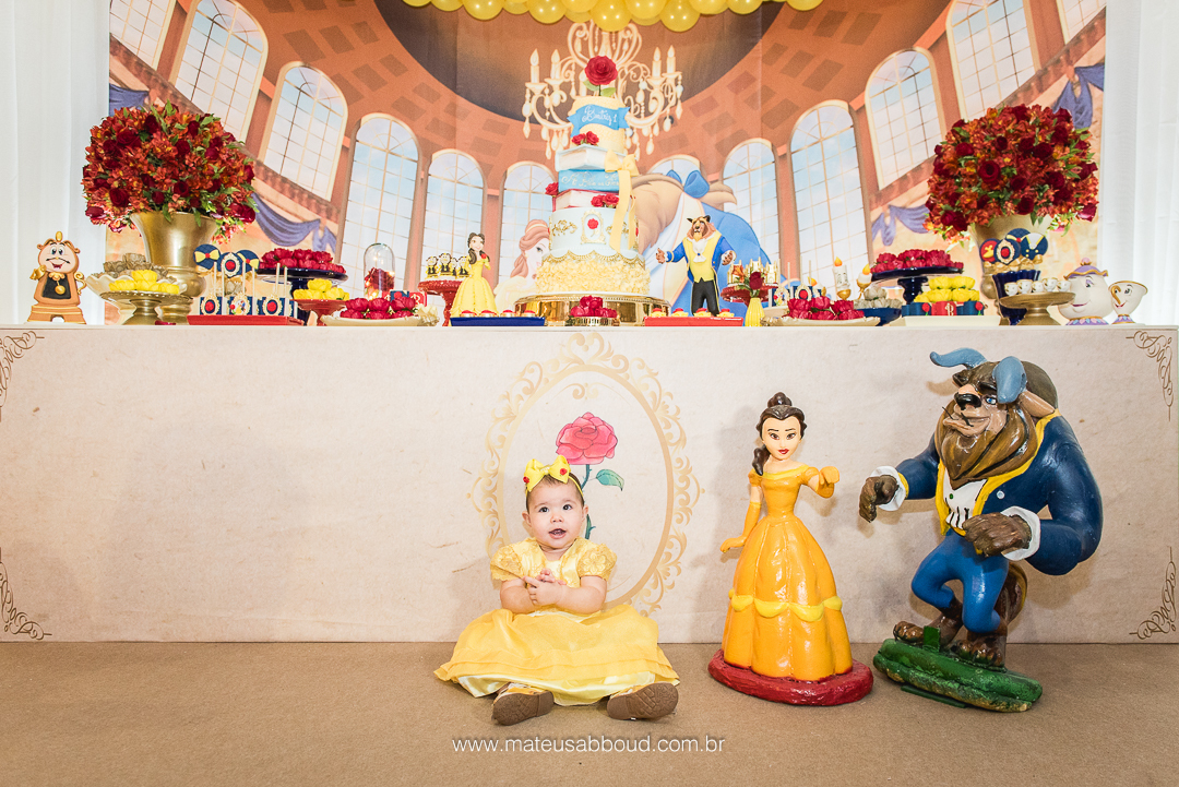 Aniversariante de 1 ano sentada no chão em frente a mesa do bolo e ao lado dos personagens de A Bela e a Fera.