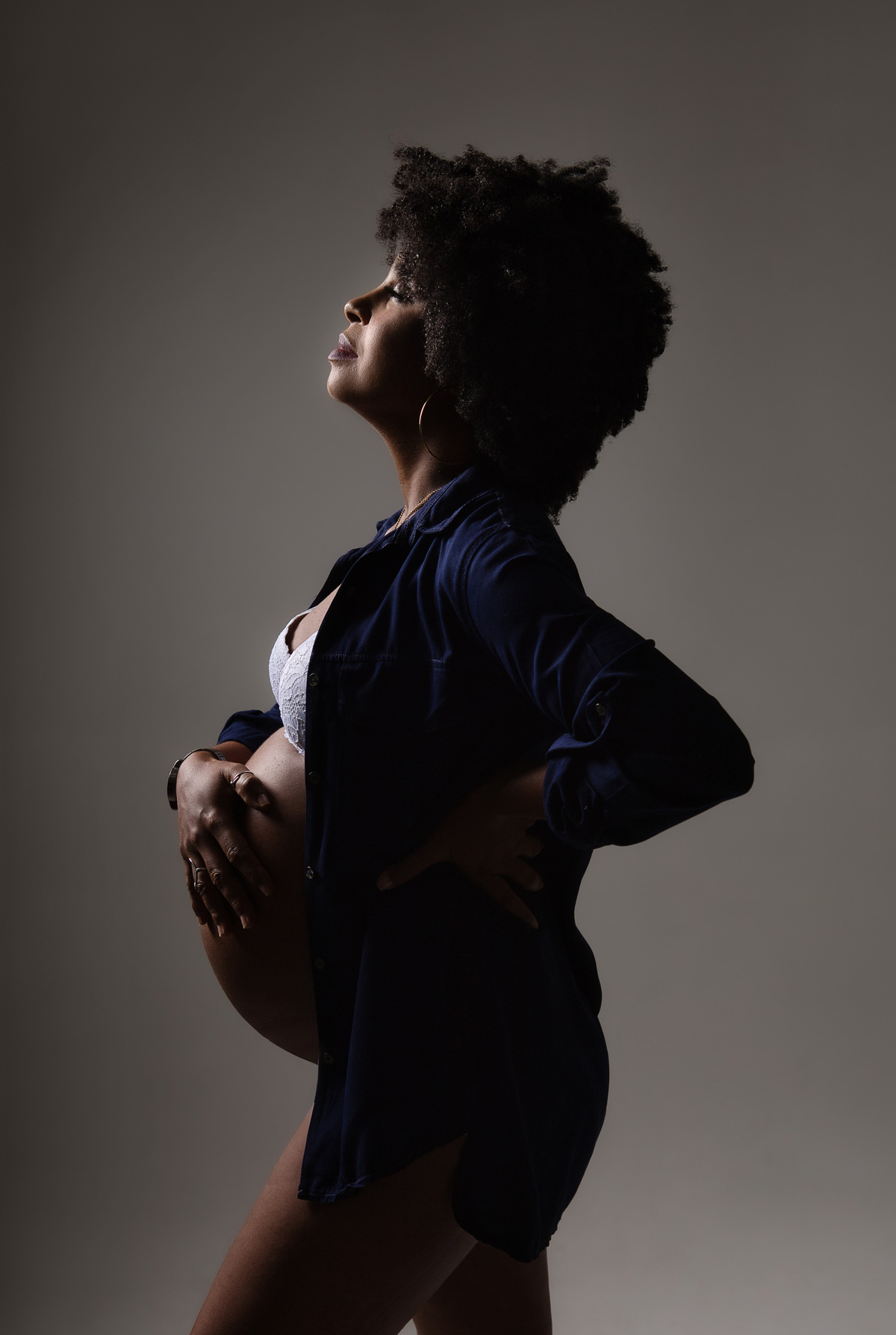 gestante com macacão e jaquetas jeans  de perfil com uma mão na barriga e outra na cintura