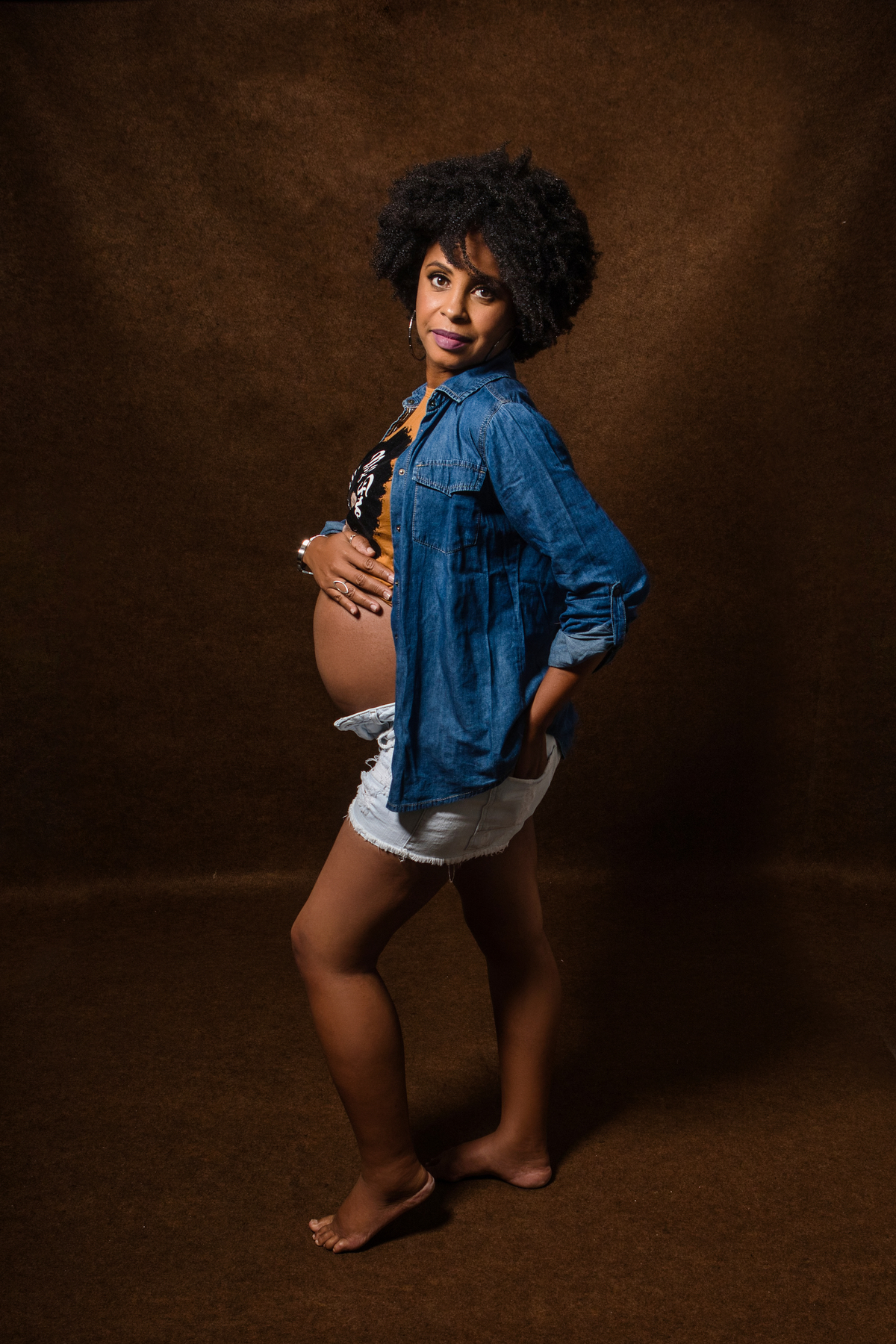 gestante com camisa, bermuda e jaqueta jeans com mão na barriga e outra na cintura