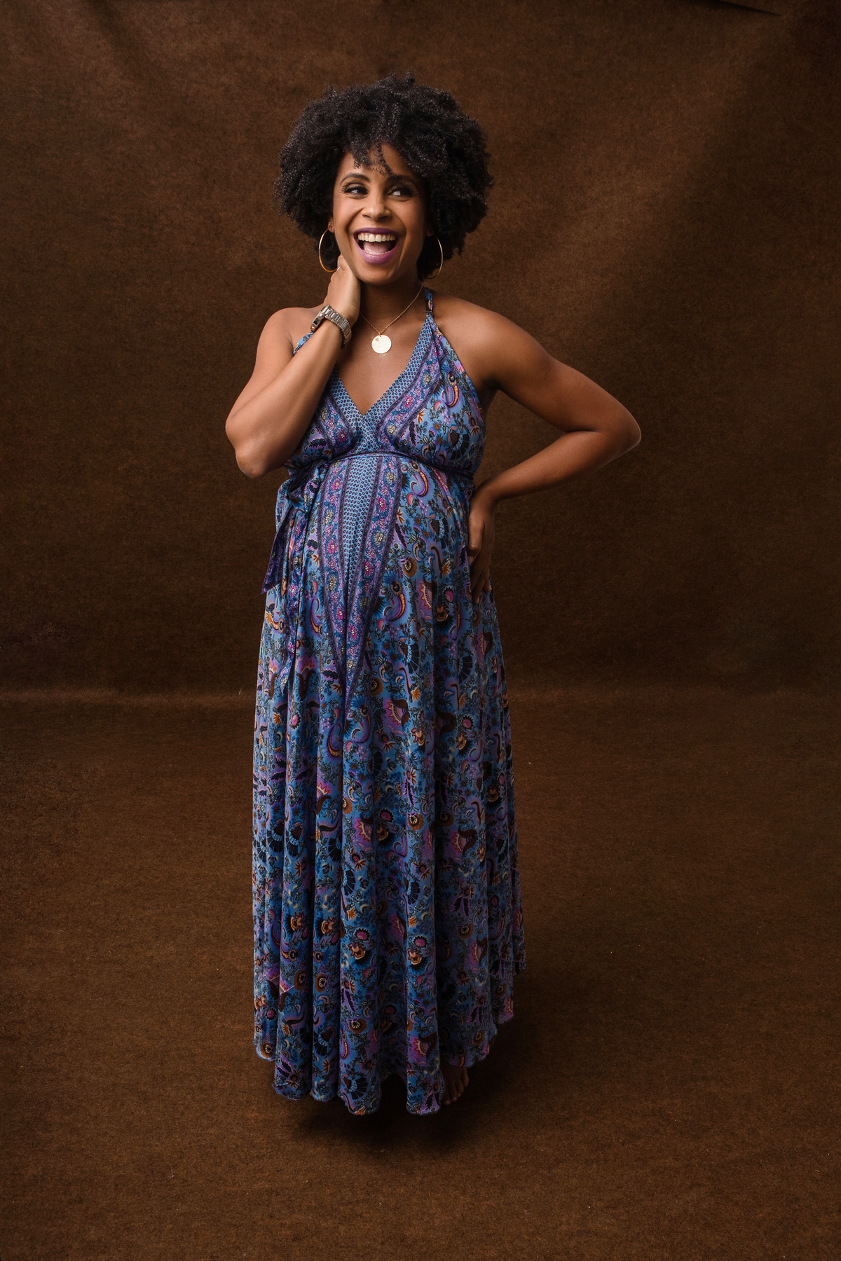 gestante sorridente de vestido longo com mão na nuca e outra na cintura 